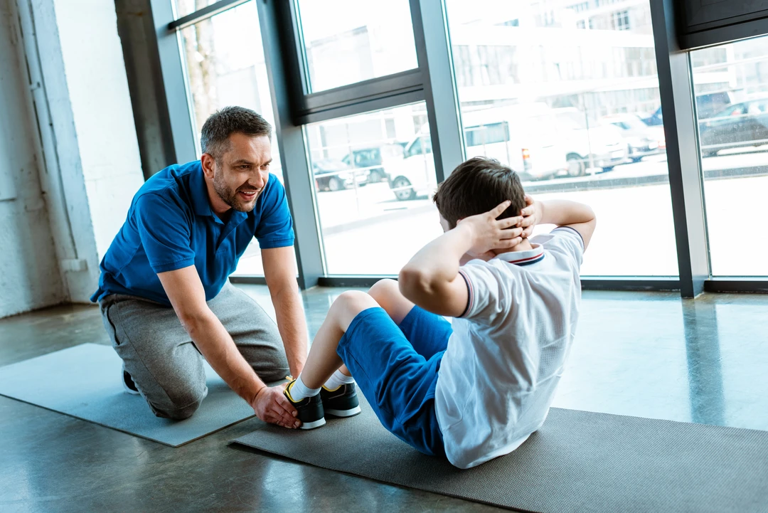 An adult holds a boy’s feet as the boy does sit-ups on a yoga mat inside a gym with large windows.